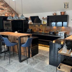 Contemporary kitchen interior, showcasing copper splashbacks and plinths, black gloss cabinets, industrial style pendant lights, and a wooden kitchen table with exposed brickwork on the walls.