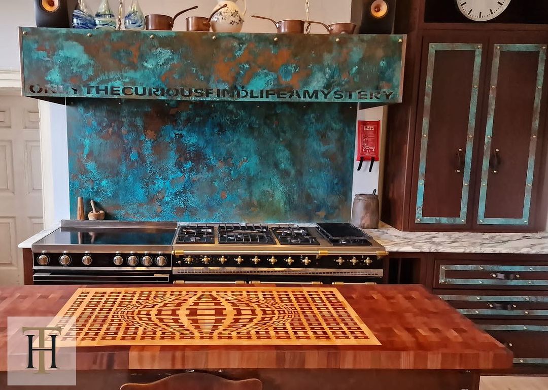 A kitchen interior showing an aged copper splashback with laser-cut lettering and a matching copper cooker hood.